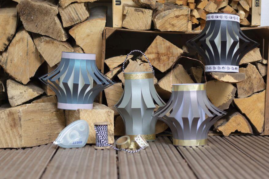 Vier selbstgebastelte Laternen aus Papier stehen aufgereiht vor einem Holzstapel.