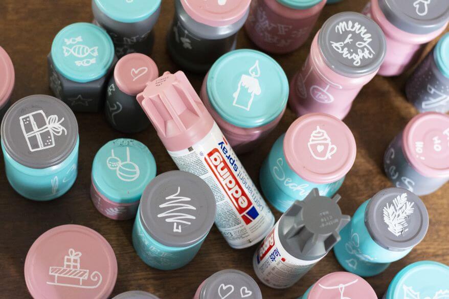 Blick von oben auf einen Upcycling-Adventskalender aus leeren Marmeladengläsern: Gläser und Deckel sind mit Sprühfarbe von Edding in grau, rosa und türkis eingefärbt mit Hilfe von Glanzlackmarkern von Edding mit kleinen, weihnachtlichen Zeichnungen versehen.