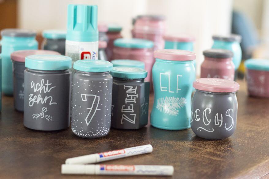 Mehrere Gläser eines Upcycling-Adventskalenders aus leeren Marmeladen- und Einmachgläsern stehen aufgereiht auf einem Tisch. Zu sehen sind verschiedene Handletteringzüge und Illustrationen auf den Gläsern.
