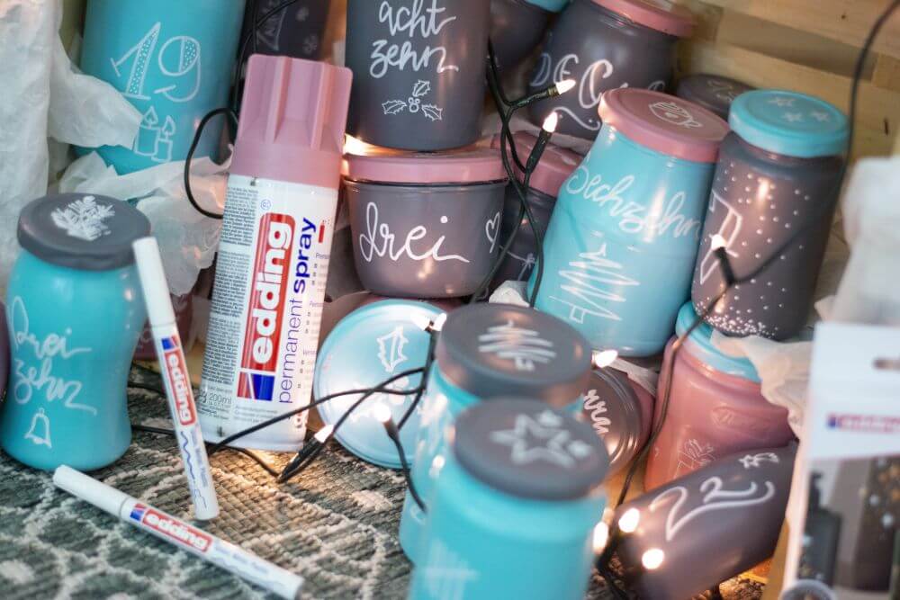 Nahaufnahme eines Upcycling-Adventskalenders aus leeren Marmeladen- und Einmachgläsern, der mit Sprühfabre und Glanzlackmarkern von Edding gestaltet wurde.