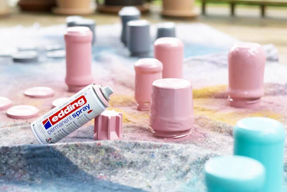 Leere Marmeladengläser, die mit Sprühfarbe von Edding in den Farben rosa, grau und türkis angesprüht wurden, stehen auf einer Malerdecke, daneben steht eine Sprühdose in rosa.