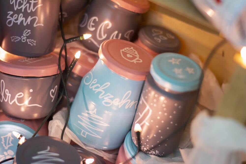 Detailaufnahme eines Upcycling-Adventskalenders aus leeren Marmeladen- und Einmachgläsern: In Handlettering ist "sechzehn" auf ein Glas geschrieben, drumherum stehen weitere Gläser und ist eine Lichterkette drapiert.