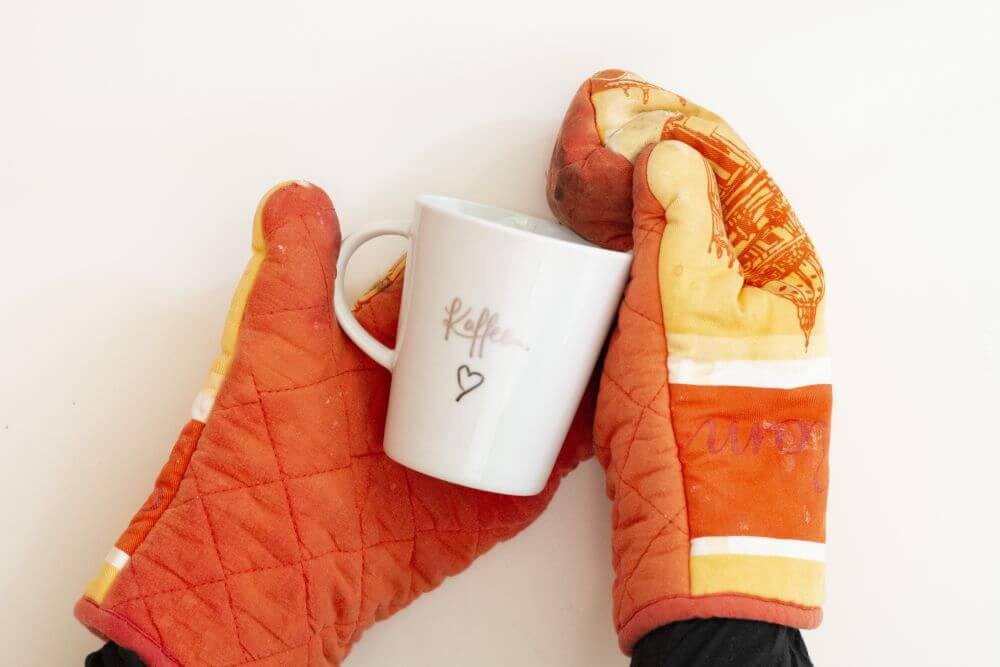 Eine Tasse, die mit Metallic-Pens von Faber Castell belettert wurde, liegt in zwei Händen, die zwei Ofenhandschuhe in orange-gelb tragen.