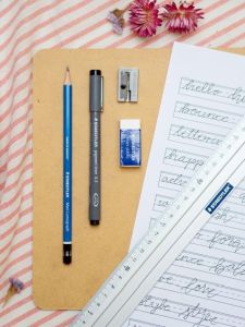 Auf einer rosa-weiß gestreiften Decke liegt ein Holz-Klemmbrett. Darauf befindet sich ein Bounce Lettering Übungsblatt, sowie alle Materialien, die man zum Lettern braucht: Bleistift, Pigment Liner, Radiergummi, Spitzer und Lineal der Marke Staedtler. Foto: Johanna Rundel