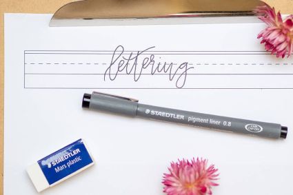 Auf einem Klemmbrett aus Holz klemmt ein Papier mit Hilfslinien zum Lettern. In den Zeilen steht das Wort „lettering“ im Bounce Lettering Stil. Pigment Liner und Radiergummi der Firma Staedtler liegen darunter. Zwei rosa Trockenblumen lockern das Bild auf. Foto: Johanna Rundel