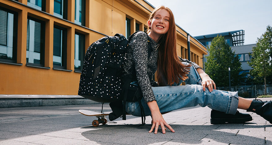 Ein Mädchen sitzt auf einem Skateboard und lächelt in die Kamera. Auf dem Rücklen trägt sie einen Schulrucksack der Marke satch.