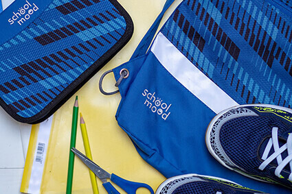 Federmäppchen und Turnbeutel der Marke SchoolMood liegen zusammen mit Heften, Schnellheftern, Stiften und Turnschuhen auf einem weißen Untergrund ausgebreitet. Alles ist in Blau- und Gelbtönen gehalten. Foto: Johanna Rundel
