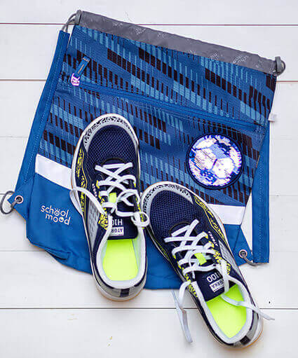 Ein blauer Turnbeutel der Marke School-Mood liegt flach auf einem weißen Untergrund. Darauf stehen ein Paar farblich passende Turnschuhe. Ein Fussball-Klett-Patch aus schillernden Wendepailletten ist das Highlight des Sportsbags. Foto: Johanna Rundel