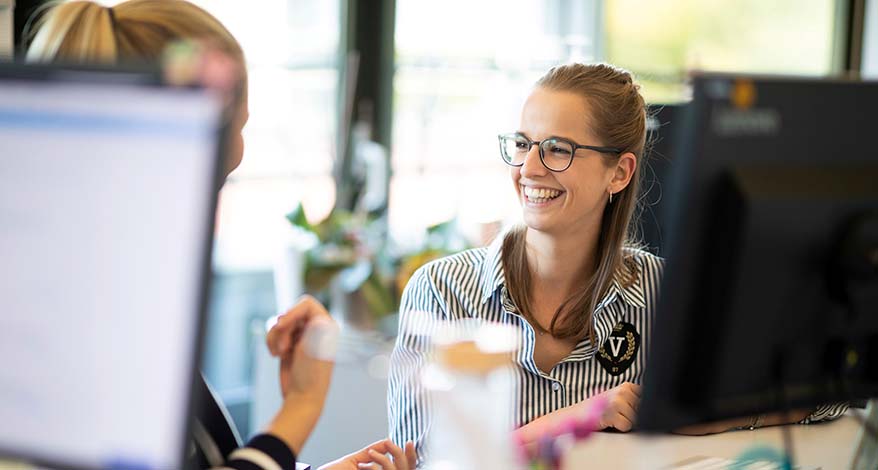 Zwei junge Frauen sitzen an einem Schreibtisch und unterhalten sich angeregt.