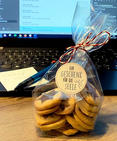 Eine Tüte mit Keksen steht vor einem aufgeklappten Laptop, auf dem ein Notizzettel und ein Kugelschreiber liegen. Auf der Tüte ist ein Designsticker der Marke Avery Zweckform aufgeklebt, auf dem steht "Ein Geschenk für die Seele"