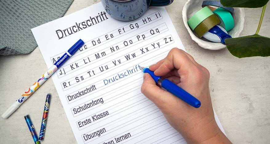 Auf einem grauen Untergrund liegt ein Vorlageblatt für ein Druckbuchstaben-Alphabet. Eine Hand füllt eine Zeile der Vorlage mit dem Combino Schreiblernfüller von Pelikan aus. Drumherum liegen ein Tintenlöscher, Tintenpatronen und eine Schale mit Washitapes. Außerdem sind eine Kaffeetasse und eine Pflanze im Bild zu sehen. Foto: Johanna Rundel