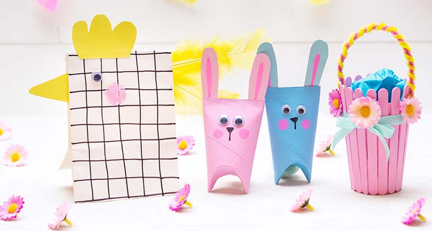 Ein rosafarbener und ein hellblauer Osterhase stehen vor einem weißen Hintergrund. Rechts davon steht ein selbstgebasteltes Osterkörbchen, aus einem Pappbecher und Bastelhölzern. Links davon steht ein Huhn, das aus einer Papiertüte gebastelt wurde.