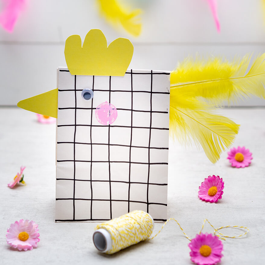 Aus einer Papiertüte wurde ein Huhn gebastelt, es steht vor einem weißen Hintergrund. Die gelben Schwanzfedern sind von Heyda. Drumherum liegen künstliche Streublumen von Knorr-Prandell. Fotos: Johanna Rundel