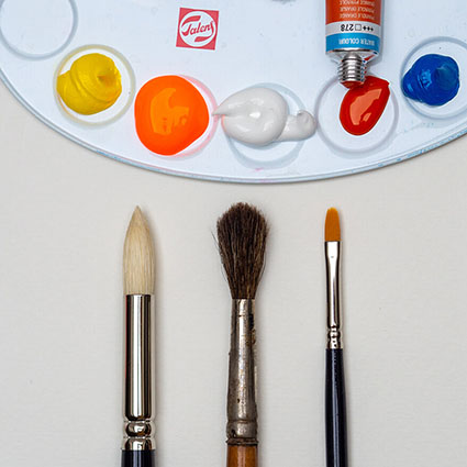 Auf einem hellen Untergrund liegen drei Pinsel. Ein Borstpinsel, ein Haarpinsel und ein Synthetikfaser-Pinsel. Man sieht nur die obere Hälfte. Oberhalb der Pinsel liegt eine Malpalette mit unterschiedlichen Farben in den Mulden. Foto: Johanna Rundel