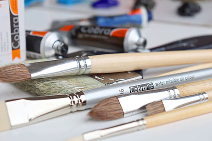 Auf einem weißen Papier liegen verschiedene Pinsel von Royal Talens. Es sind nur die vorderen Hälften zu sehen. Im Hintergrund liegen Farbtuben und eine Malpalette.