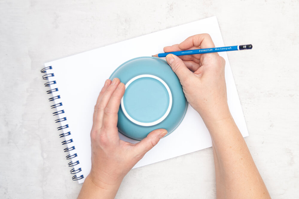Jemand zeichnet den Umriss einer hellblauen Schüssel mit einem Bleistift der Marke Staedtler auf ein Malblock. Foto: Johanna Rundel