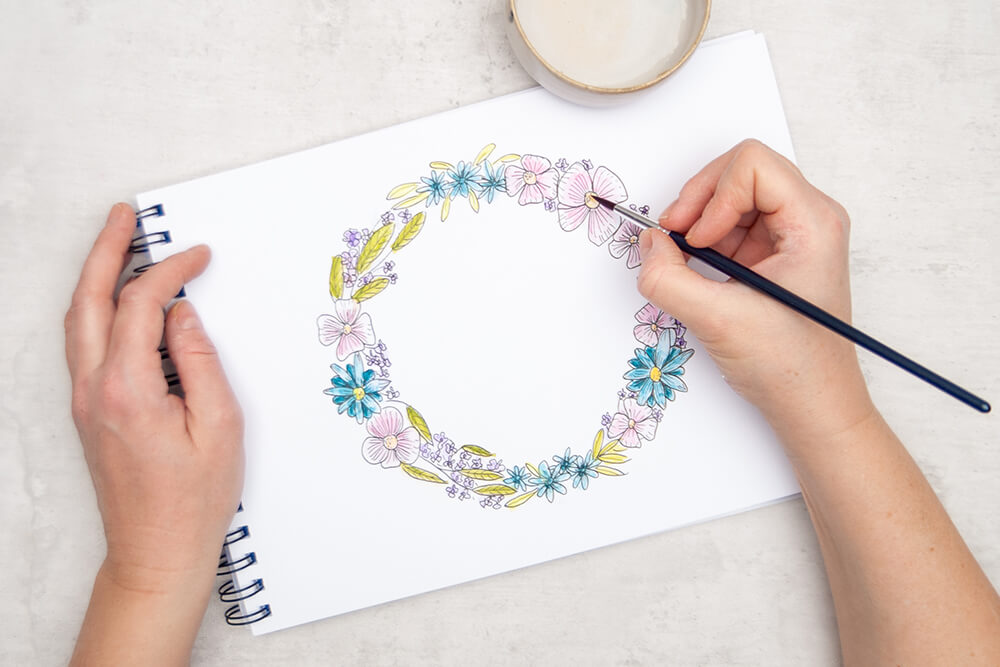 Jemand aquarelliert die Blüten und Blätter eines mit wasservermalbaren Buntstiften von Staedtler kolorierten Blumenkranzes mit Wasser und einem Pinsel. Foto: Johanna Rundel