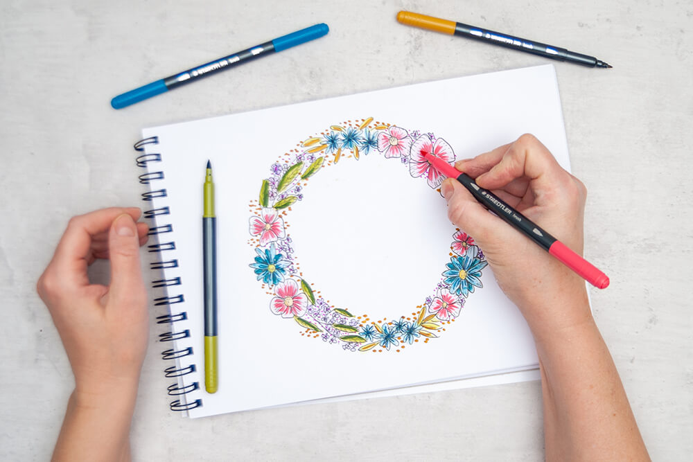 Jemand setzt mit den Double-ended watercolour brush pens von Staedtler Akzente in einem gezeichneten und kolorierten Blumenkranz. Foto: Johanna Rundel