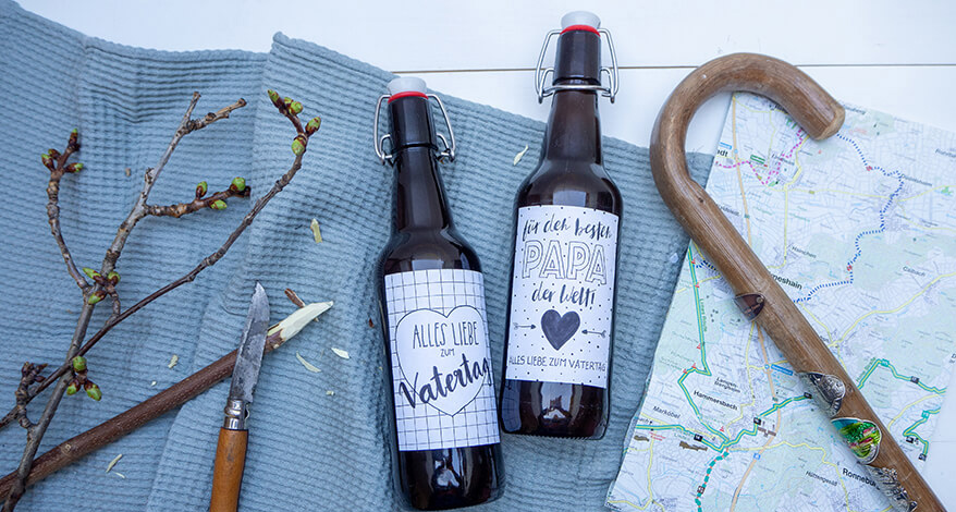 Auf einem Tisch liegt ein blaues Leinentuch, darauf liegen zwei Bierflschen, die mit einem selbst gestaltetetn Etikett beklebt sind. Daneben liegt ein Wanderstock, eine Wanderkarte, ein Zweig und ein Messer zum Schnitzen. Foto: Johanna Rundel. .