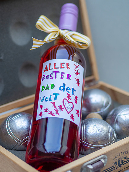 In einem Boule-Koffer aus Holz liegt auf den Boule-Kugeln eine Flasche Roséwein. Auf der Flasche klebt ein von Kinderhand selbstgestaltetes Etikett von Avery Zweckform mit Glückwünschen zum Vatertag. Foto: Johanna Rundel