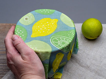 Eine Hand schmiegt ein gemustertes Bienenwachstuch um eine kleine Schüssel. Daneben liegt eine grüne Limette. Foto: Johanna Rundel