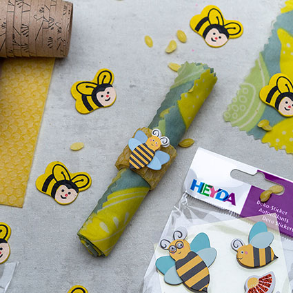 Auf einem hellgrauen Untergrund liegt ein eingerolltes Bienenwachstuch, das mit einem Bienensticker verziert wurde. Lustige Streudeko in Form von kleinen Holzbienen von Knorr-Prandell liegen darum verteilt. In der rechten unteren Ecke liegt ein Stickerbogen der Firma Heyda mit 3-D Bienenaufklebern. Foto: Johanna Rundel