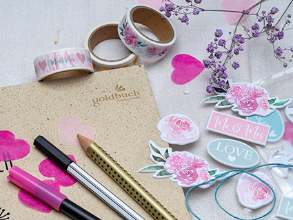 Auf und um ein hellbraunes Hochzeitsgästebuch von Goldbuch liegen verschiedene Stifte, Washi Tapes, 3-D Aufkleber und echte Blumen verstreut. Foto: Johanna Rundel