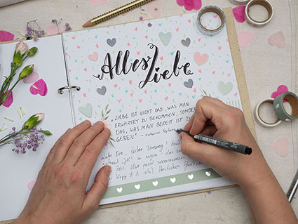 Auf einem hellen Untergrund liegt ein aufgeschlagenes Hochzeitsgästebuch von Goldbuch. In großen Letteringbuchstaben steht dort „Alles Liebe“ geschrieben. Jemand schreibt gerade Glückwünsche hinein. Die Seite ist kreativ mit Aufklebern, Washi Tape und bunten Stiften gestaltet. Die benutzen Materialien liegen um das Buch verstreut. Überall sind Papierherzen und Blümchen verstreut. Foto: Johanna Rundel