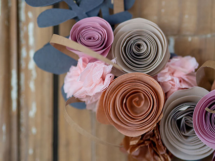 An einer rustikalen Holztür hängt ein goldener Reif mit verschiedenen Papierblumen und -blättern. Die verwendeten Papiere sind alle von der Firma Artoz. Es ist nur das untere linke Viertel des Kranzes im Detail zu sehen. Foto: Johanna Rundel