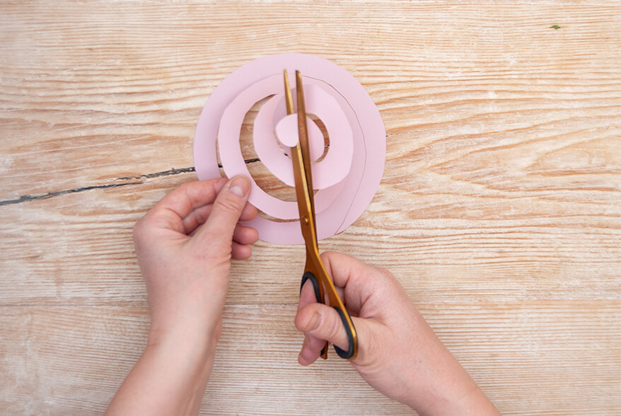 Auf einer hellen Holzoberfläche schneidet jemand eine Spirale aus einem Kreis rosafarbenen Tonpapiers von Artoz. Foto: Johanna Rundel