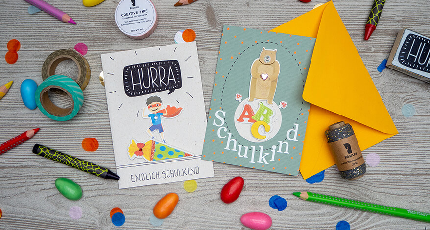 Auf einem hellgrauen Holzuntergrund liegen eine wasserblaue Grußkarte, auf die ein brauner Bär, die Buchstaben ABC und das Wort Schulkind mit Aufklebern der Firma Rössler geklebt ist. Daneben liegt eine weiße Karte, mit einem gestempelten "Hurra", einem Superhelden-Kind, das auf einer Schultüte steht und der Spruch "Endlich Schulkind" drauf. Drumherum liegen buntes Konfetti, eine Garnspule, Washitape und verschiedene Stifte verteilt.
