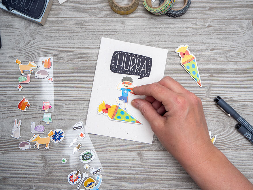 Auf einem hellgrauen Holzuntergrund liegt eine weiße Grußkarte der Marke Rössler. Darauf klebt jemand Sticker mit Schulmotiven. Über den Stickern steht "Hurra".