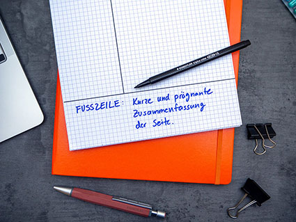 Auf einem dunklen Untergrund liegt ein orangefarbenes Notizbuch, darauf ein karierter Notizblock. Auf dem Block ist die Aufteilung für die Cornell Methode gezeichnet. Der untere Bereich für die Zusammenfassung ist mit blauem Stift farblich hervorgehoben. Auf dem Block liegt ein Fasermaler der Marke Staedtler. Darunter ein Kugelschreiber.