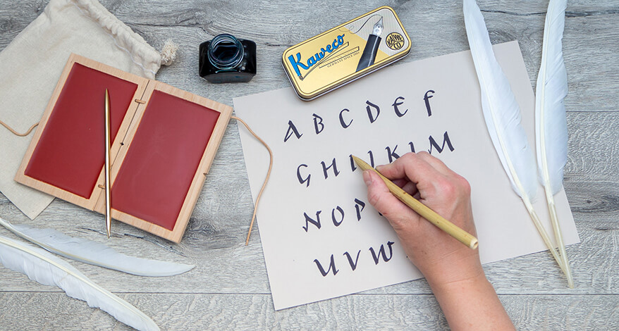 Auf einem rustikalen Holzuntergrund liegt ein Papier, auf dem mit römischer Schrift ein Alphabet geschrieben steht. Daneben liegt ein kleiner Jutebeutel, auf dem eine römische Wachstafel mit rotem Wachs und ein Griffel aus Bronze liegen. Oberhalb davon ist ein Tintenfass und eine Blechdose mit einem modernen Füllfederhalter von Kaweco zu sehen. Foto: Johanna Rundel