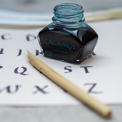 Auf einem rustikalen Holzuntergrund liegt ein Papier, auf dem mit römischer Schrift ein Alphabet geschrieben steht. Auf dem Papier steht ein dunkles CALAMVS-Tintenfass von Aratrum. Ein angespitztes Schilfrohr (Calamus) liegt daneben. Foto: Johanna Rundel