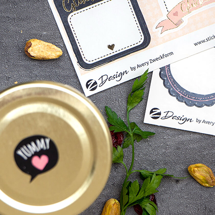 Auf einem dunkeln Untergrund steht ein kleines Marmeladenglas mit goldenem Deckel. Auf dem Deckel klebt ein schwarzer Aufkleber in Form einer Sprechblase mit der Aufschrift "Yummy". Im Hintergrund liegen zwei Bögen Design-Aufkleber von Avery-Zweckform, sowie Pistazien und getrocknete Cranberrys. Foto: Johanna Rundel