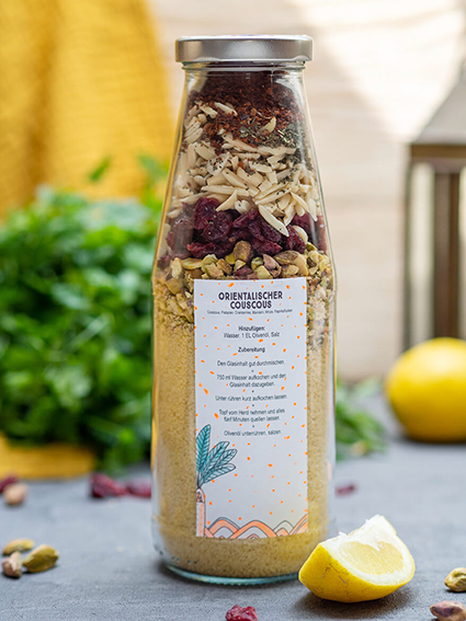 Auf einem dunkeln Untergrund steht eine Glasflasche mit silbernem Deckel. Darin ist eine Couscous-Mischung – die Zutaten sind dekorativ übereinandergeschichtet. Auf der Flasche klebt ein weißer Aufkleber von Avery Zweckform mit den Zubereitungshinweisen. Im Hintergrund liegt ein Bund Petersilie, im Vordergrund und um das Glas herum sind Pistazien, Zitronenspalten und getrocknete Cranberrys verteilt. Foto: Johanna Rundel