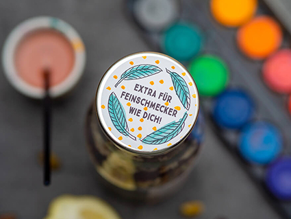 Auf einem dunkeln Untergrund steht eine Glasflasche mit silbernem Deckel. Auf der Flasche klebt ein Aufkleber von Avery Zweckform. Darauf steht: "Extra für Feinschmecker wie dich". Der Aufkleber wurde mit Wasserfarben coloriert. Der Wasserfarbkasten sowie Pinsel und Wasserbehälter sind im Hintergrund verschwommen sichtbar. Foto: Johanna Rundel