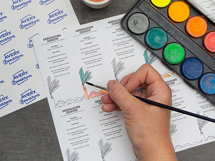 Jemand malt mit einem Pinsel und bunten Wasserfarben einen schwarz-weiß bedruckten Bogen Klebeetiketten von Avery Zweckform farbig an. Der Wasserfarbkasten, der Wasserbehälter und ein weiterer Bogen Etiketten sind ebenfalls im Bild zu sehen. Foto: Johanna Rundel
