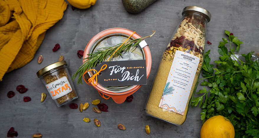 Auf einem dunkeln Untergrund liegen von links nach rechts: Ein kleines Marmeladenglas mit goldenem Deckel und der Aufschrift Zatar. Ein Weck-Glas mit eingelegten Oliven, einem Rosmarinzweig und einem Etikett von Avery Zweckform auf dem "Für Dich" steht. Eine Glasflasche, die mit einer Couscous-Mischung gefüllt ist – auf der Flasche klebt ein Etikett mit den Zubereitungshinweisen. Darum verteilt liegen ein Bund Petersilie, ein Mörserstab, ein gelbes Handtuch, Zitronen, Pistazien und getrocknete Cranberrys verteilt. Foto: Johanna Rundel