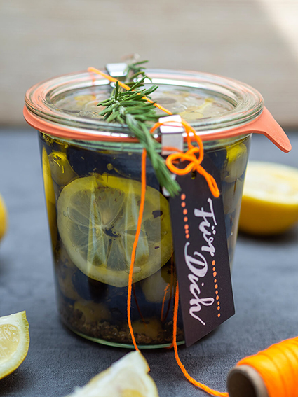 Auf einem dunkeln Untergrund steht ein Weck-Glas mit eingelegten Oliven. Mit einem Bäckergarn in neonorange ist ein Rosmarinzweig und ein schwarzes Etikett von Avery Zweckform mit der Aufschrift "Für Dich" daran gebunden. Drumherum sind Zitronenspalten dekoriert. Foto: Johanna Rundel