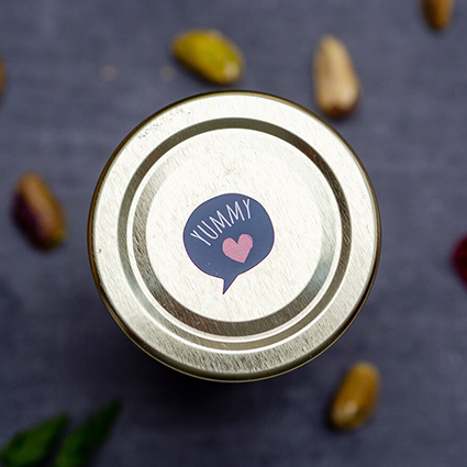 Auf einem dunkeln Untergrund steht ein kleines Marmeladenglas mit goldenem Deckel. Auf dem Deckel klebt ein schwarzer Aufkleber in Form einer Sprechblase mit der Aufschrift "Yummy". Pistazien und getrocknete Cranberrys sind unscharf im Hintergrund zu sehen. Foto: Johanna Rundel