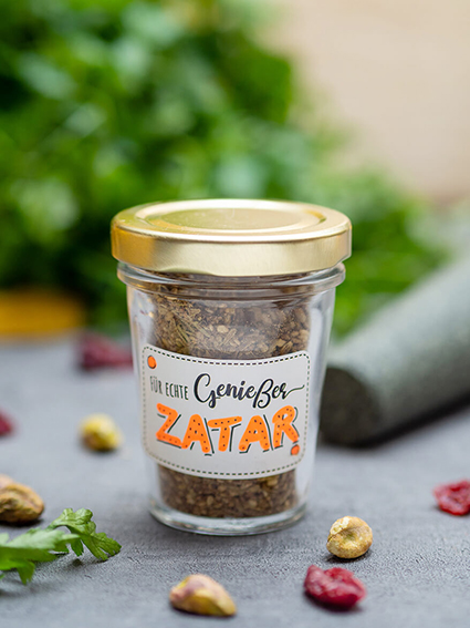 Auf einem dunklen Untergrund steht ein kleines Marmeladenglas mit goldenem Deckel. Darauf ist ein weißer Aufkleber von Avery-Zweckform mit der Aufschrift "Für echte Genießer – Zatar". Im Hintergrund liegt ein Bund Petersilie und ein Mörserstab. Im Vordergrund und um das Glas herum sind Pistazien und getrocknete Cranberrys verteilt. Foto: Johanna Rundel