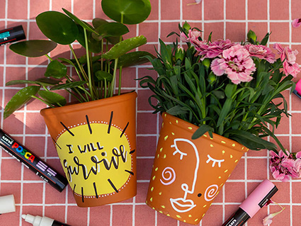 Auf einer rosa-weiß karierten Tischdecke liegen zwei Blumentöpfe aus Ton, die mit bunten Markern angemalt wurden. Auf dem linken Topf steht „I will survive“ auf. Er ist mit einer Pilea-Pflanze bestückt. Auf dem rechten Topf ist in Line-Art ein Frauengesicht gezeichnet. Im Topf befindet sich eine pinke Nelke. Um die beiden Töpfe herum sind verschiedene Marker von Posca verteilt. Foto: Johanna Rundel