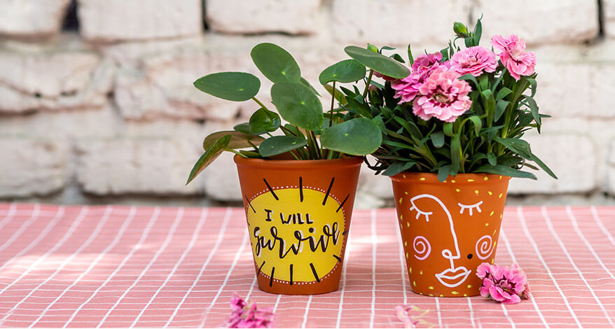 Auf einer rosa-weiß karierten Tischdecke stehen zwei Blumentöpfe aus Ton, die mit bunten Markern von Posca angemalt wurden. Auf dem linken Topf steht „I will survive“. Er ist mit einer Pilea-Pflanze bestückt. Auf dem rechten Topf ist in Line-Art ein Frauengesicht gezeichnet. Im Topf befindet sich eine pinke Nelke. Im Hintergrund ist eine weiße Backsteinwand zu sehen. Foto: Johanna Rundel