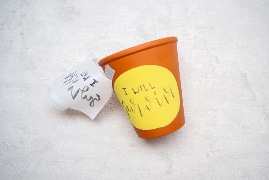 Auf einen Blumentopf aus Ton wurde mit Markern von Posca ein gelbes Oval gemalt. Darauf steht mit Bleistift der Satz „I will survive“. Seitlich am Blumentopf hängt, mit Washitape befestigt, ein Stück Butterbrotpapier mit der Vorlage für das Lettering. Der Untergrund ist hell. Foto: Johanna Rundel