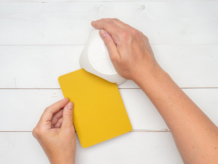 Jemand rundet mit einem 4-in-1-Stanzer von Artoz die Ecken einer gelben Blanko Klappkarte ab. Der Untergrund ist weiß. Foto: Johanna Rundel