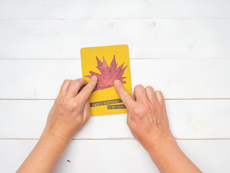 Jemand klebt zwei getrocknete Herbstblätter auf eine gelbe Blanko Klappkarte von Artoz, auf die „Happy Birthday“ gestempelt wurde. Der Untergrund ist weiß. Foto: Johanna Rundel