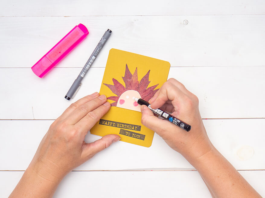 Jemand malt ein Gesicht auf einen rosa Halbkreis aus Tonkarton, der auf zwei getrocknete Herbstblätter geklebt wurde. Das Motiv klebt auf einer gelben Blanko Klappkarte von Artoz, auf welche "Happy Birthday" gestempelt wurde. Der Untergrund ist weiß. Foto: Johanna Rundel