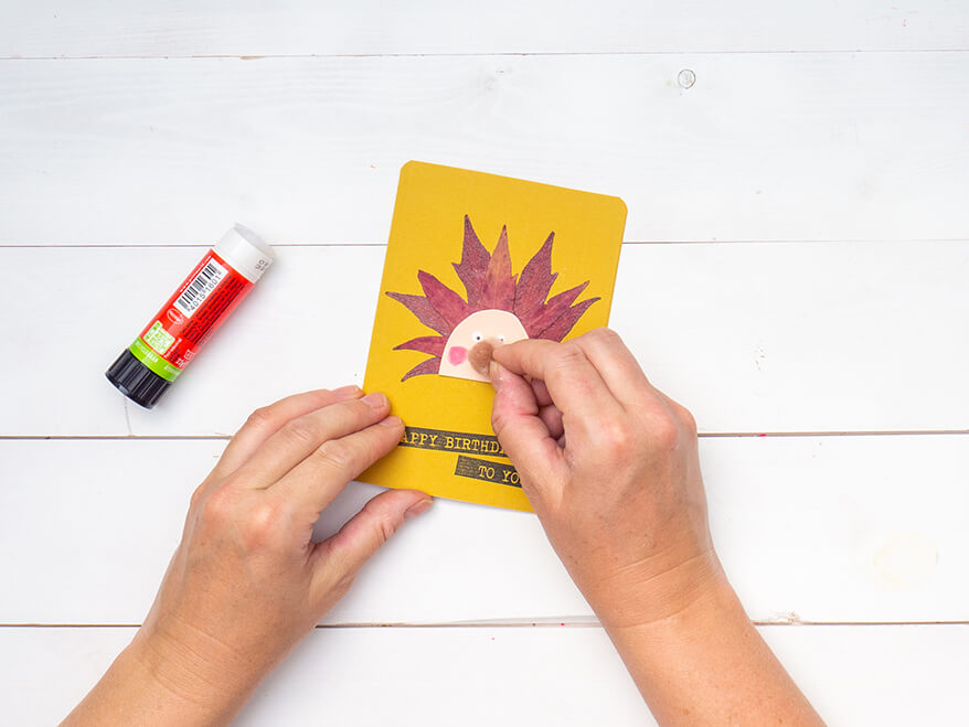 Jemand klebt einen braunen Mini-Pompon auf einen rosa Halbkreis aus Tonkarton mit Gesicht. Der Untergrund ist weiß. Foto: Johanna Rundel